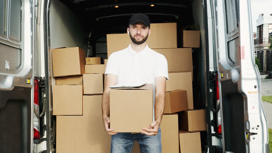 A man wearing a white shirt, dark baseball cap, and jeans is standing inside the open rear of a large moving van, which is filled with numerous uniformly-sized cardboard boxes stacked neatly on top of each other and along the sides. The man is holding a single box securely with both hands, and a clipboard is resting on top of the box. The van is parked outside on a paved surface in a residential area, with houses and greenery visible in the background. Natural daylight illuminates the scene, highlighting the clean, organized interior of the van used for home relocation or furniture transport, as part of a professional moving service by manwithvanelephantandcastle.com. The environment suggests an active loading process, with the boxes prepared for transit to a new location, exemplifying efficient packing and loading techniques for house removals and relocation services.