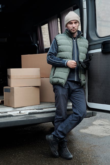 A man wearing a white shirt, dark baseball cap, and jeans is standing inside the open rear of a large moving van, which is filled with numerous uniformly-sized cardboard boxes stacked neatly on top of each other and along the sides. The man is holding a single box securely with both hands, and a clipboard is resting on top of the box. The van is parked outside on a paved surface in a residential area, with houses and greenery visible in the background. Natural daylight illuminates the scene, highlighting the clean, organized interior of the van used for home relocation or furniture transport, as part of a professional moving service by manwithvanelephantandcastle.com. The environment suggests an active loading process, with the boxes prepared for transit to a new location, exemplifying efficient packing and loading techniques for house removals and relocation services.