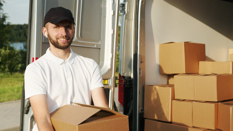 A man with a beard, wearing a black cap and a white polo shirt, is seen during a home relocation process involving furniture transport and packing. He is standing outside, next to an open moving van, which is parked on a driveway with a lush green background and blue sky visible. Inside the van, several cardboard boxes are arranged on the floor and stacked neatly, indicating an active loading process. The man is holding a cardboard box in his hands, preparing to load it into the van, with additional boxes seen behind him, some in clear view and others partially obscured. The presence of packing materials and the outdoor environment suggests the final stages of packing and loading for a house move, carried out by [COMPANY_NAME], emphasizing professional furniture transport and moving logistics for the relocation to or from the Walworth Road area as detailed in the Elephant and Castle removals guide for Walworth Road moves.