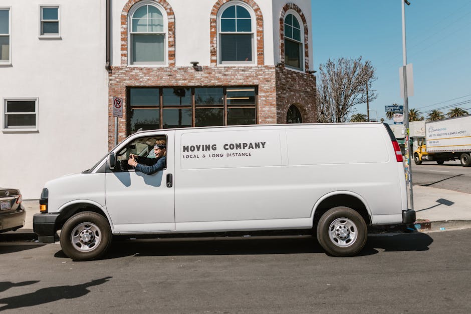 A white moving van with the text 'MOVING COMPANY LOCAL & LONG DISTANCE' printed on its side is parked on a city street in front of a brick building with large windows. The van's driver, wearing sunglasses and a dark jacket, is seated in the cab, holding the steering wheel and looking forward. The pavement beside the van shows some shadows cast by nearby objects, and other vehicles are visible in the background. The scene depicts a professional furniture transport and home relocation process, typical of services provided by [COMPANY_NAME], with the van ready for loading or unloading in a move along Walworth Road. The urban environment features a blue sky and some trees, indicating daytime, and the area appears prepared for the logistics involved in furniture transport and packing during a house removal.