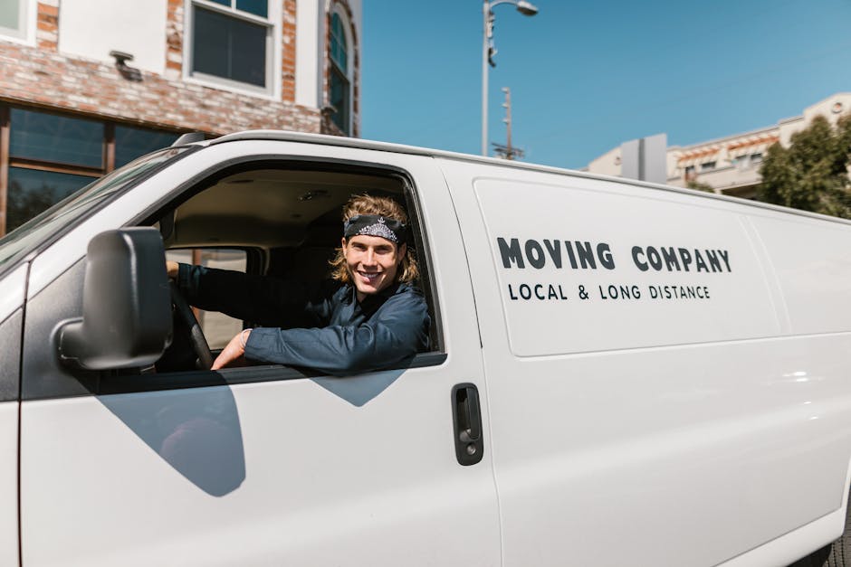 A man with a beard, wearing a black cap and a white polo shirt, is seen during a home relocation process involving furniture transport and packing. He is standing outside, next to an open moving van, which is parked on a driveway with a lush green background and blue sky visible. Inside the van, several cardboard boxes are arranged on the floor and stacked neatly, indicating an active loading process. The man is holding a cardboard box in his hands, preparing to load it into the van, with additional boxes seen behind him, some in clear view and others partially obscured. The presence of packing materials and the outdoor environment suggests the final stages of packing and loading for a house move, carried out by [COMPANY_NAME], emphasizing professional furniture transport and moving logistics for the relocation to or from the Walworth Road area as detailed in the Elephant and Castle removals guide for Walworth Road moves.