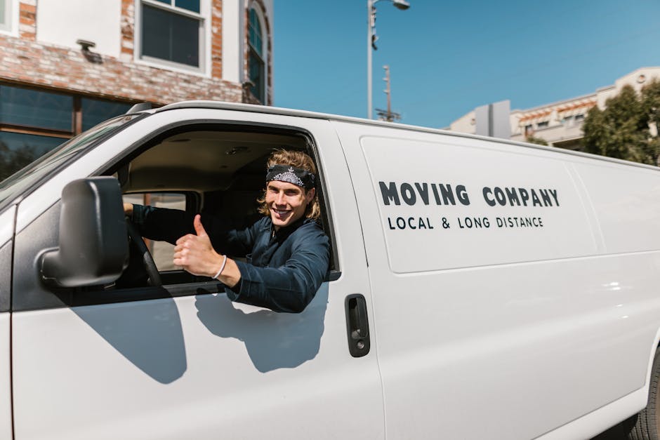 A young male mover with long hair, wearing a patterned headband and casual dark clothing, is sitting in the driver's seat of a white moving van labeled 'MOVING COMPANY LOCAL & LONG DISTANCE.' He is smiling and giving a thumbs-up through the open window, indicating a positive attitude during the home relocation process. The van is parked outdoors on a street with modern buildings, windows, and a clear blue sky in the background. The scene reflects a professional movers' team preparing for furniture transport, packing, and loading in a typical house removal or local move setting, with the focus on ensuring safe and efficient moving logistics. The vehicle includes equipment such as side mirrors and part of the sliding side door, typical of vans used for removals carried out by companies like [COMPANY_NAME], supporting effective packing and moving services.