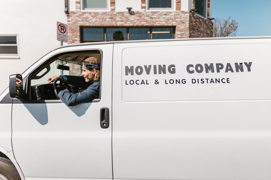 A white moving van parked on a street in front of a modern brick building with large windows, featuring the text 'MOVING COMPANY LOCAL & LONG DISTANCE' printed on the side in bold black letters. Inside the van, a professional mover with long blonde hair, wearing a navy blue jacket and a black headband, is sitting in the driver’s seat, preparing for a home relocation. The background includes a clear blue sky, a no-parking sign, and a paved sidewalk. The scene captures the loading process and the vehicle ready for furniture transport or packing and moving services offered by the company, aligning with house removals in the SE1 area.