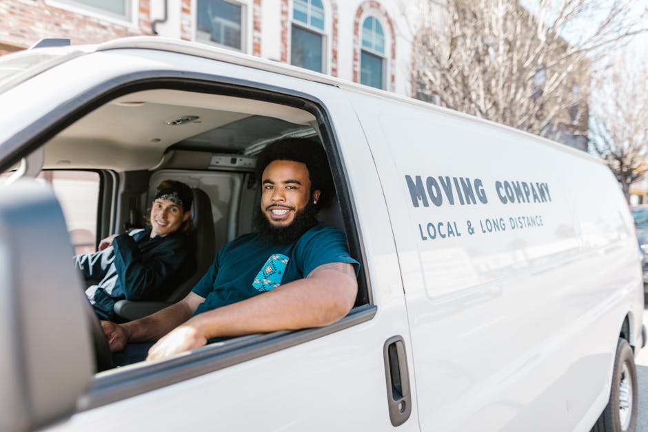 Inside a white moving van marked with the words 'Moving Company, Local & Long Distance,' two male movers are seated in the front cabin. The driver, a man with a dark hair and a beard, is smiling and looking at the camera. The passenger, a man with dark hair, is also smiling and looking towards the camera. Visible from the open driver's side window are packed cardboard boxes, some wrapped in plastic, placed on the passenger seat and in the back of the van, indicating preparations for home relocation. The van is parked outdoors near a residential area with trees and multi-storey buildings visible in the background. The space appears well-lit, with natural daylight illuminating the interior and exterior environment, and the movers seem ready for a furniture transport or packing and moving process with the assistance of [COMPANY_NAME].
