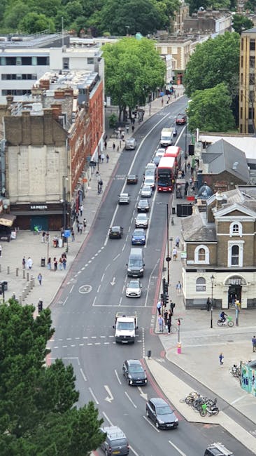 An aerial view of Newington Causeway in Elephant and Castle showing a busy urban street with multiple lanes of traffic, including cars, vans, and buses moving in both directions. The street is lined with a mix of modern and historic buildings, some with brick facades and others with flat rooftops. Several pedestrians are walking along the sidewalks, and a few bicycles are parked near the curb. Large, leafy green trees are visible along the pavements, providing shade to the pedestrians. The scene also features a loading and unloading area where a man with a van, associated with a house removal service, is positioned to facilitate furniture transport and home relocation tasks, employing packing materials like cardboard boxes and protective blankets. The overall environment suggests a typical city centre setting focused on logistical activities related to moving and transport services, with evident movement of vehicles and activity around the property for efficient house removals and relocation efforts.