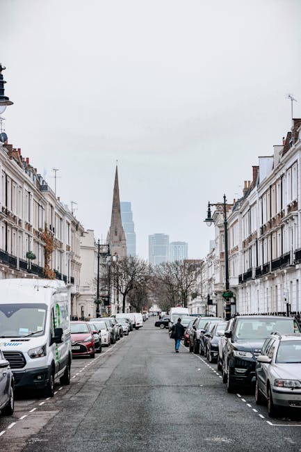 A view of a residential street in central London, with rows of white Victorian-style terraced houses on either side, each featuring black wrought-iron balconies and large windows. Parked cars line both sides of the street, including a white van on the left and various sedans and hatchbacks on the right. In the foreground, a person wearing dark clothing is seen loading or unloading a box or piece of furniture into or from a vehicle parked near the curb. The street is paved with asphalt, and a few leafless trees are visible along the sidewalks. In the background, the spire of a church or historic building rises above the rooftops, with modern skyscrapers visible further behind, indicating an urban environment typical of a London home relocation or furniture transport scenario. The scene is overcast, with diffused natural light illuminating the area, emphasizing the typical moving or packing process handled by companies like [COMPANY_NAME], offering professional removals services in the Elephant and Castle area.