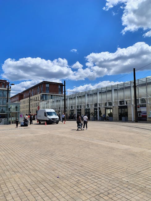A spacious outdoor area in front of a modern building with large glass windows and metal framing, situated near Elephant and Castle station. The scene shows a bright day with a clear blue sky and scat